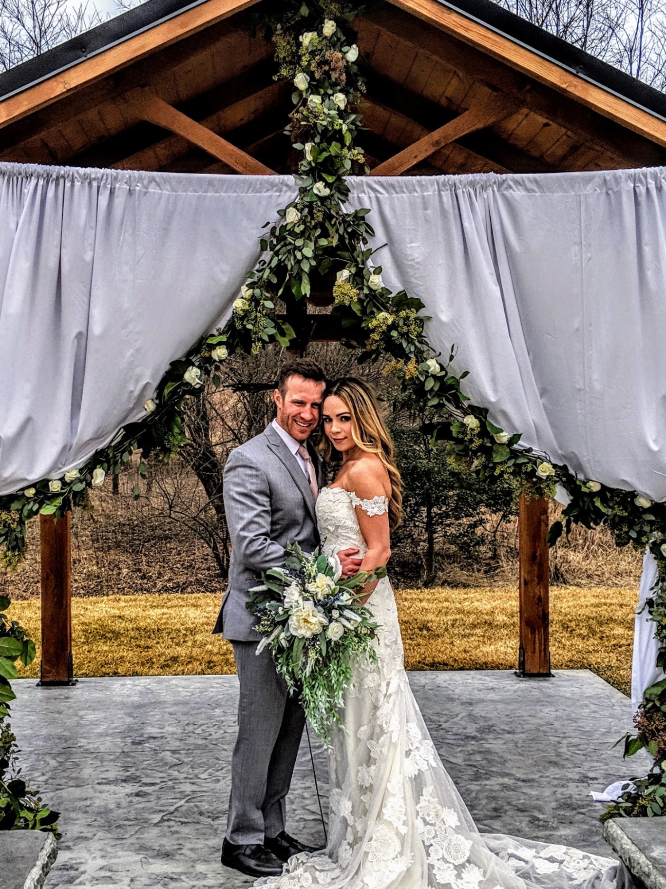 Couple at the pavilion arch Bride and groom under the pavilion draped in grey fabric with a white rose and eucalyptus arch