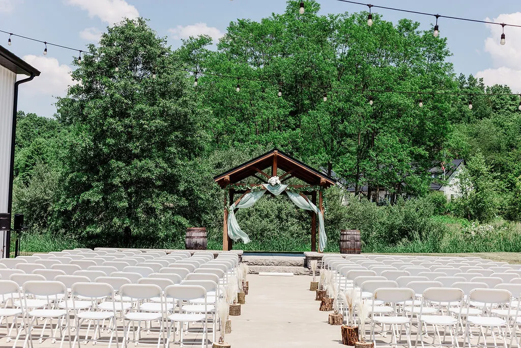 Outdoor ceremony setup with white chairs, wooden arbor and sage draping