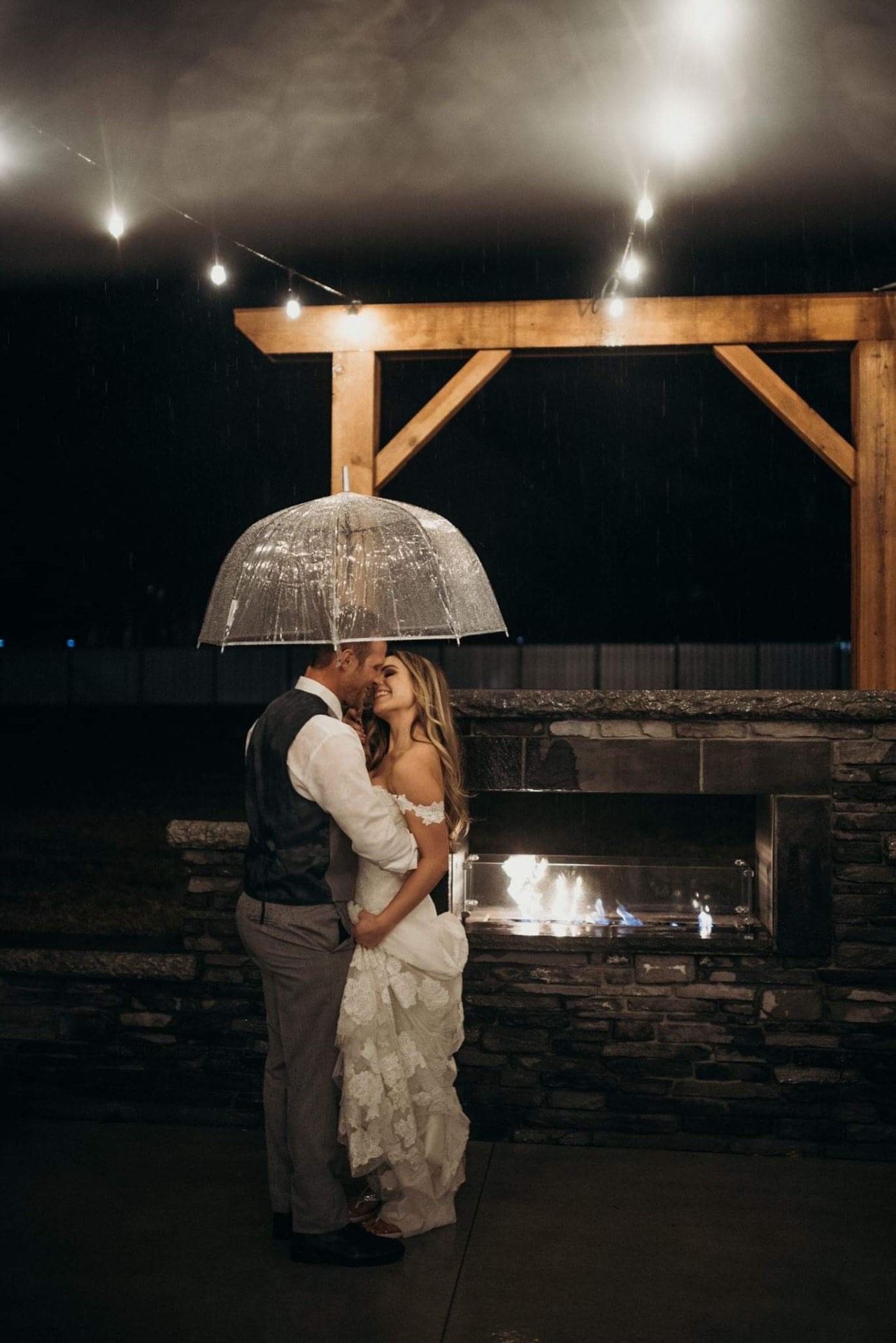Rainy night by the fireplace Couple embracing under a clear umbrella beside the lit outdoor fireplace at night in the rain
