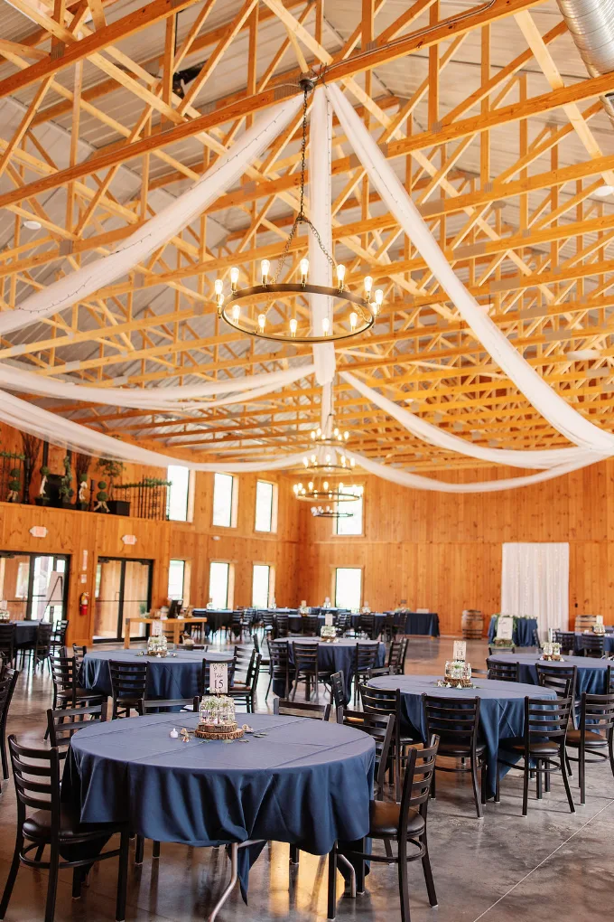 Round guest tables with navy linens beneath timber trusses inside the barn