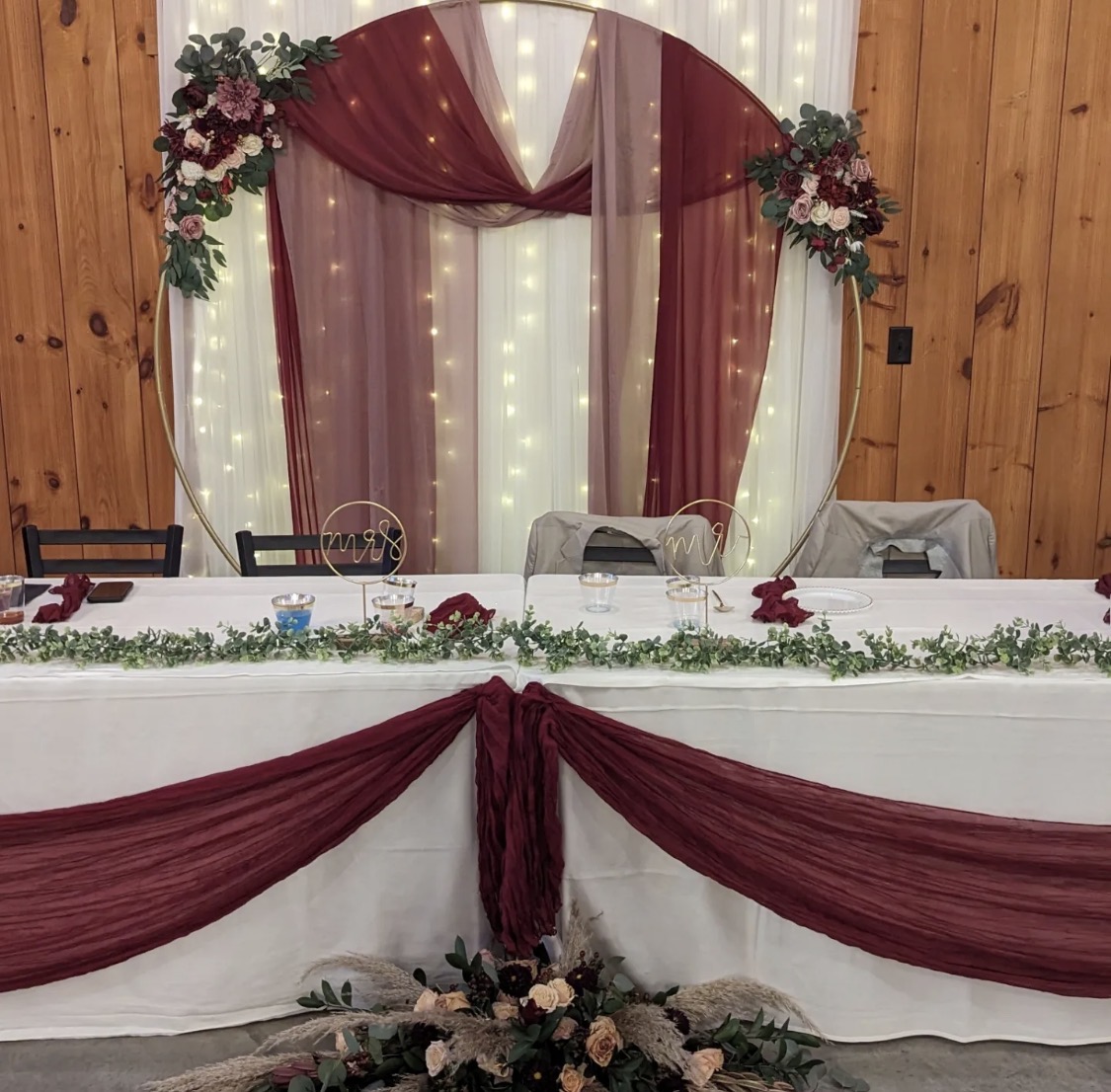 Head table with burgundy draping and a gold hoop arch inside the barn